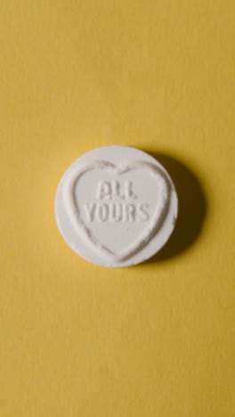 Vertical-Video-Close-Up-Of-Vertical-Video-Of-Hand-Holding-Heart-Candy-With-First-Love-Message-On-Yellow-Background-1